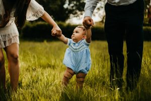 One year old baby walking in grass field holding his parents hands